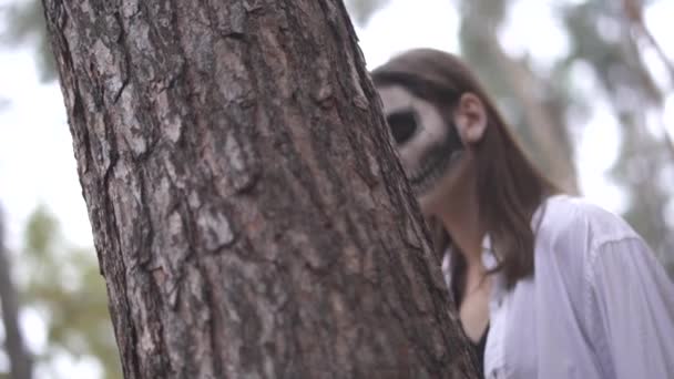 Halloween. Chica con un cuchillo y maquillaje de Halloween escondido detrás de un árbol — Vídeos de Stock