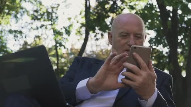 Un anciano está mirando un teléfono inteligente mientras está sentado en un banco en el parque — Vídeo de stock