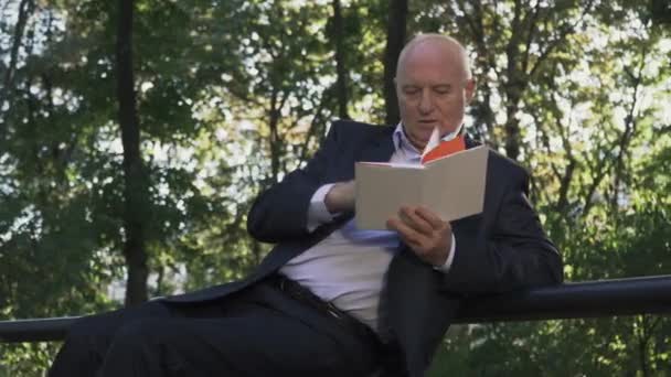 El viejo está leyendo un pequeño libro en el parque. — Vídeo de stock