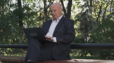 Yaşlı adam ile laptop dizlerinin üstünde oturuyor. Olgun işadamı parkta çalışıyor.