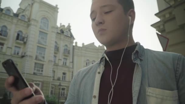 Молодой человек в наушниках слушает музыку и смотрит в телефон — стоковое видео