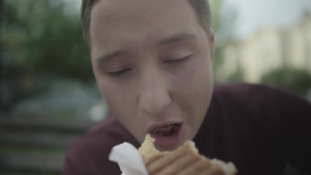 Jonge man eten sandwich buitenshuis. Gezicht close-up — Stockvideo