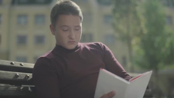 Здивоване обличчя хлопця, який читає книгу на відкритому повітрі — стокове відео