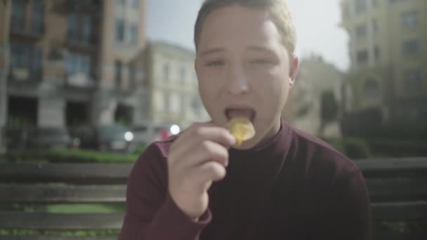 Chico come patatas fritas con placer mientras está sentado en un banco — Vídeos de Stock