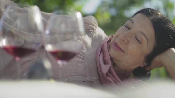 Porträt einer reifen Frau, die mit einem Glas Wein auf dem Gras im Garten liegt — Stockvideo