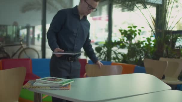 Junger Mann mit Netbook kommt und sitzt am Tisch in moderner Bibliothek — Stockvideo