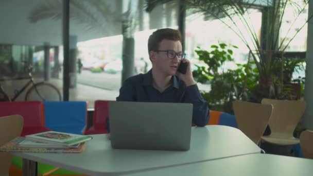 El tipo de las gafas hablando por teléfono mientras está sentado en un portátil — Vídeo de stock