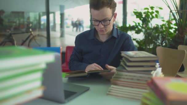 Hombre guapo está estudiando con muchos libros sobre la mesa en la biblioteca — Vídeos de Stock