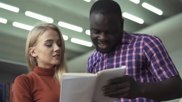 Mulher europeia e homem afro-americano lendo livro na biblioteca — Vídeo de Stock