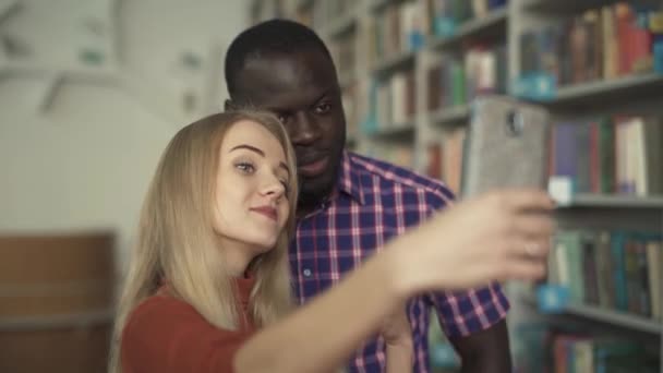 Hombre afroamericano y señora europea se toman selfie en la biblioteca — Vídeos de Stock