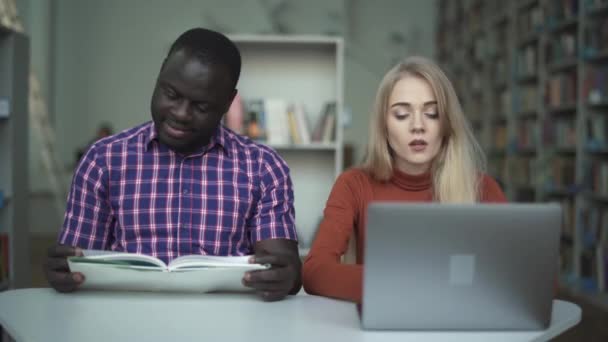 Chica cansada con un portátil y su amigo afroamericano leyendo un libro — Vídeo de stock