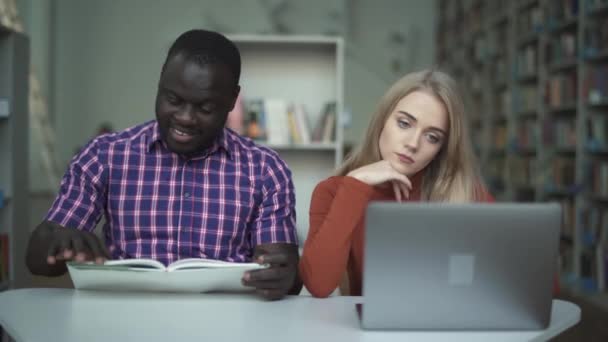 Linda chica cansada con un portátil y su amigo afroamericano leyendo un libro — Vídeo de stock