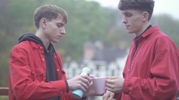 Молодой парень наливает чай из термоса в чашку другу Друзья проводят время на свежем воздухе вместе Мужская дружба — стоковое видео