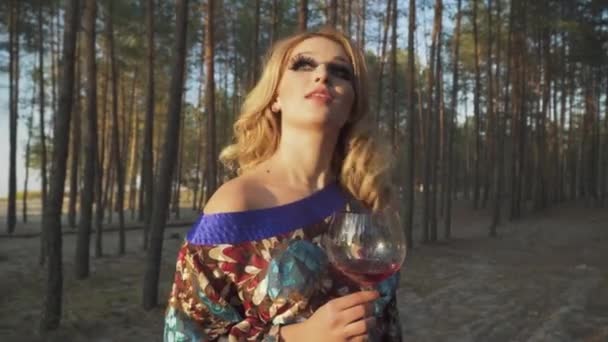 Preciosa mujer de pie en el bosque con copa de vino Señora con pestañas falsas sonrisas Chica en vestido de verano con hombro desnudo bebe vino al aire libre Rubia con ojos verdes y pestañas largas — Vídeos de Stock