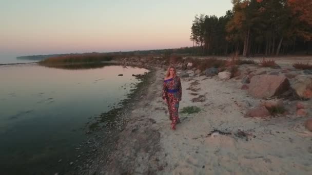 Menina bonita em um vestido de verão com um ombro nu caminha ao longo da costa do lago Mulher bonito com cabelo loiro caminha ao ar livre Menina em vestido de verão com ombro nu bebe vinho ao ar livre Tiro — Vídeo de Stock