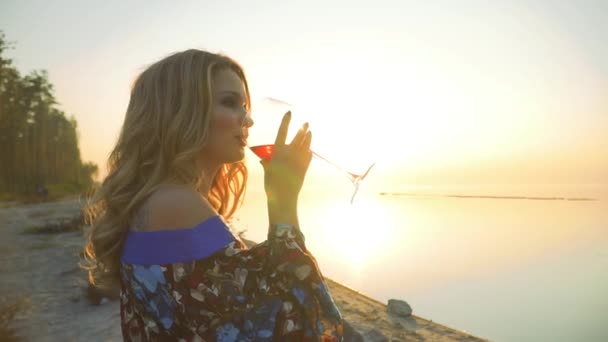 Красива дівчина сидить на березі дивовижного озера на сонці п'є вино з келиха Дівчина в літній сукні з голими плечима напої вино на відкритому повітрі Сонце в келиху вина — стокове відео