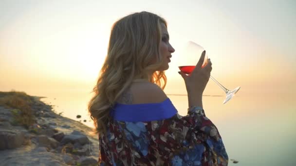 Портрет блондинки, пьющей вино на берегу моря крупным планом Леди наслаждается пейзажем в одиночестве Девушка в летнем платье с голыми плечами напитки вина на открытом воздухе Солнце в бокале вина — стоковое видео