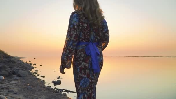 Frau geht bei Sonnenuntergang weg Frau in langem hellen Kleid spaziert am Strand — Stockvideo