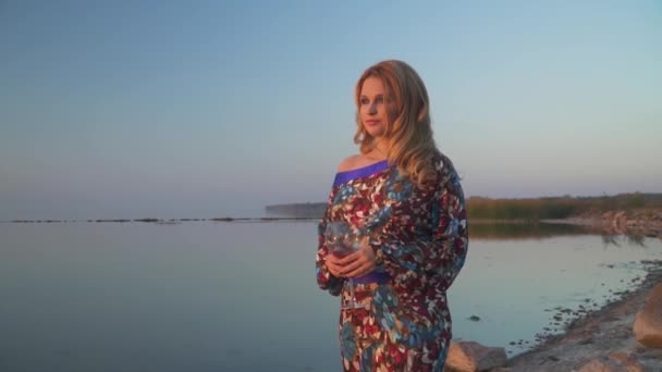 Lovely girl in a summer dress with a bare shoulder walks along the shore of the lake Cute woman walks outdoors Girl in summer dress with bare shoulder drinks wine outdoors — Stock Video