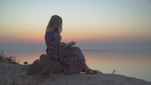 Mujer linda con el pelo rubio leyendo el libro Mujer está descansando junto al mar Chica en vestido de verano con hombro desnudo — Vídeos de Stock