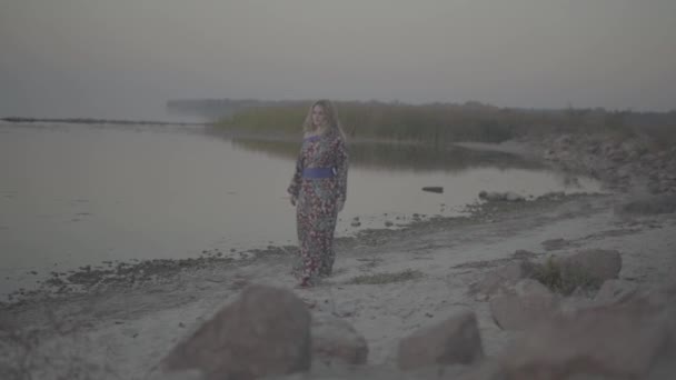 Vrouw in een zomerjurk met een blote schouder wandelingen langs de oever van het meer Cute vrouw wandelingen buiten meisje in zomerjurk met blote schouder dranken wijn buitenshuis — Stockvideo