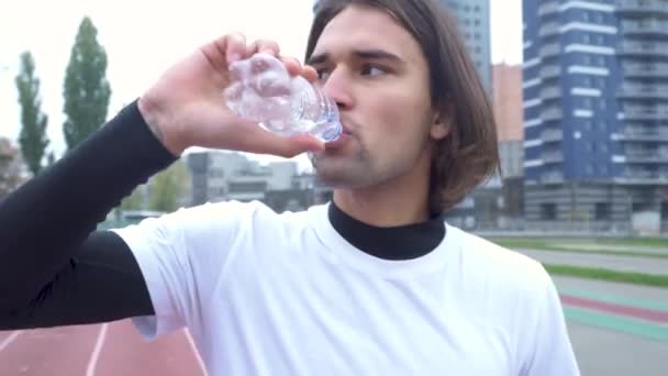 Jonge kerel in een sport shirt drinkt water op het stadion knappe man met donkere haren dranken water uit een fles na een training buitenshuis gezonde levensstijl — Stockvideo