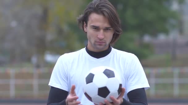 Retrato de un joven con una camiseta deportiva sosteniendo una pelota de fútbol en sus manos y mirando estrictamente a la cámara Tipo deportivo con una pelota en sus manos con una mirada descarada — Vídeo de stock