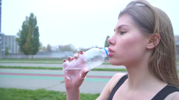 Charming girl in a sports shirt and sweat pants drinks water at the stadium Beautiful girl with dark hair drinks water from a bottle after a workout outdoors Sports girl with beautiful lips drinking — Stock Video