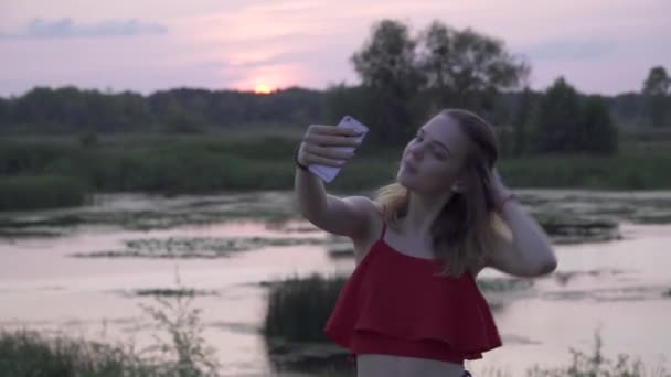 Ragazza carina posa sulla macchina fotografica telefono sullo sfondo del tramonto Giovane donna sta facendo selfie sul tramonto Femmina sta riposando all'aperto — Video Stock