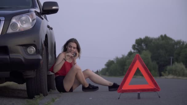 Chica molesta cerca de su coche destrozado Mujer joven está sentada cerca de coche roto Mujer es triste Mujer está llamando por teléfono móvil Problemas de coche — Vídeo de stock