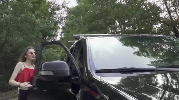 Chica delgada en camisa roja y gafas se sienta en gran coche negro Empresa de tres personas que viajan Una niña y dos amigos viajan en coche — Vídeo de stock