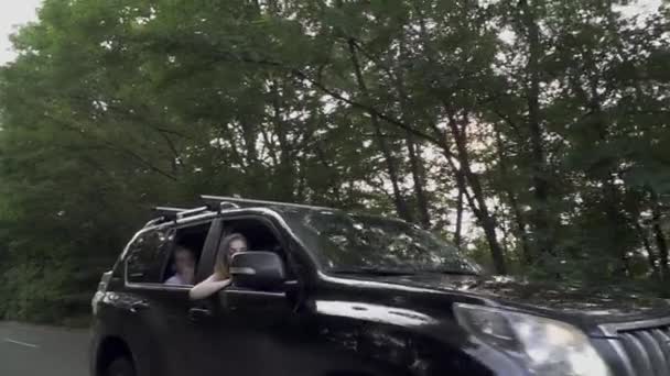 Coche negro pasea lentamente por la carretera Compañía de tres personas viajan Chica saca la cabeza por la ventana del coche — Vídeo de stock
