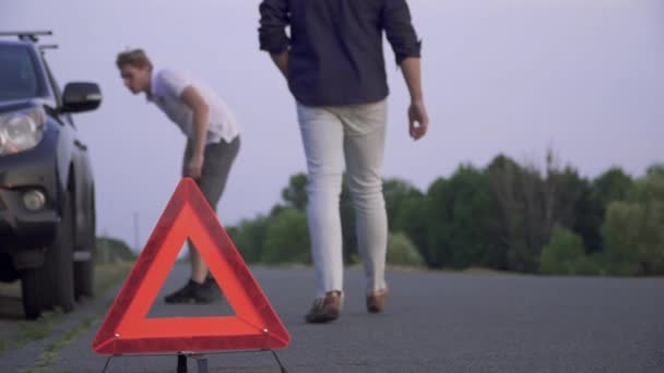 Разбитая черная машина стоит на дороге Человек в белой джинсе ставит предупреждающий треугольник на асфальт Автомобиль проблемы Фокус на переднем плане — стоковое видео