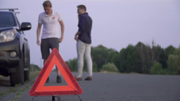 Dos amigos al lado del coche roto. Los jóvenes están molestos por la avería del coche. Un tipo pide ayuda por teléfono y el otro está mirando el volante. — Vídeo de stock