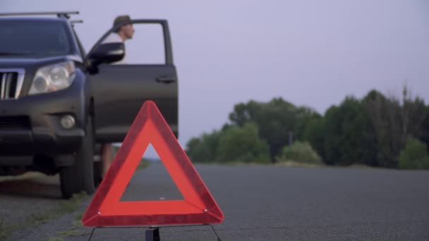 Az ember kap az autó, és megpróbálja gumiabroncs autó volt törve, a közúti figyelmeztető háromszög lassítva lábával állva aszfalt autó baj fókusz az előtérben — Stock videók