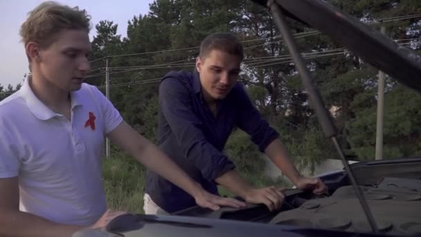 Dois jovens olham para o capô aberto de seu carro Amigos têm veículo quebrado na estrada Problemas de carro — Vídeo de Stock