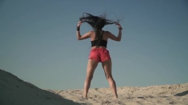 Twerk sahilde dans çekici genç kız. Tabaklanmış kız kısa kırmızı şort kumsalda onu vücut ayakta taşır. Kadın açık havada zaman harcamaktadır. Arkadan çekim