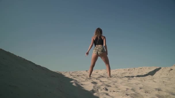 Спортсменка танцює тверк на пляжі Засмагла дівчина в коротких шортах рухає її тіло, стоячи на піску Стрілянина ззаду — стокове відео