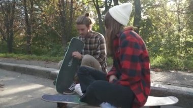 Açık havada oturan genç kaykay çift. Kız bir ekose gömlek ve beyaz şapka bir kaykay üzerinde oturan ve bir adamla konuşuyor. Adam onun kaykay ayarlama.