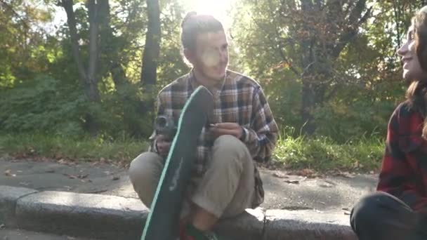 Le gars avec la fille communique assis dans le parc. Le gars ajuste une planche à roulettes en parlant à une fille . — Video
