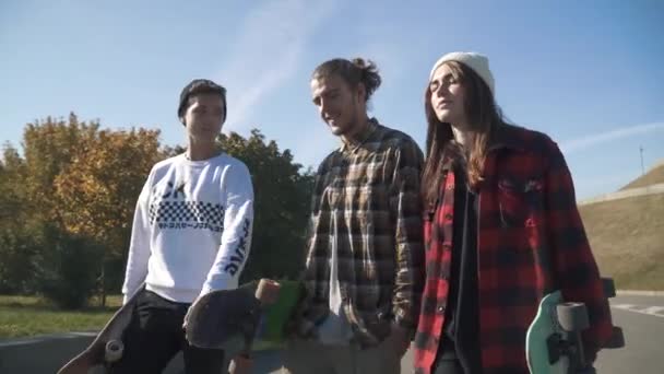Una compañía de jóvenes sonrientes llevan patinetas en sus manos mientras caminan por el parque. Los patinadores pasan tiempo al aire libre. Pasatiempos y estilo de vida . — Vídeos de Stock