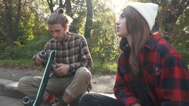 Jonge skateboarden paar zitten buiten Hipster meisje in een plaid shirt en een witte hoed praten met een jongen. Skateboarder man past zijn skateboard. Slow motion. — Stockvideo