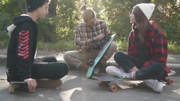 Teenagers are sitting with skateboards next to each other outdoors. Hipster friends chatting while sitting on a skateboard in the park. Young guy in a plaid shirt adjusts his skateboard. — Stock Video