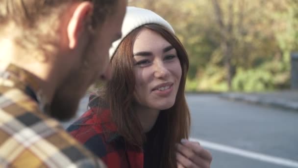 Guy y chica se comunican sentados en el parque. Chica sonriente con un sombrero blanco y camisa a cuadros hablando con un chico al aire libre . — Vídeos de Stock