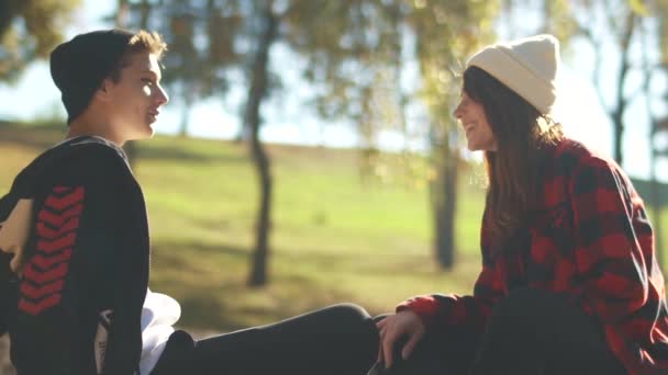 Хіпстер і дівчина спілкуються, сидячи на сонці в парку. Підлітки розслабляються на відкритому повітрі. Скейтбордистська дівчина в білому капелюсі та сорочці. Скейтбордист хлопчик в темному капелюсі і светрі — стокове відео