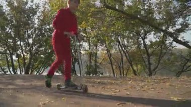 Kırmızı gömlek ve pantolon parkta bir scooter sürme içindeki küçük çocuk. Çocuk aktif açık havada zaman harcıyor. Ağır çekim.