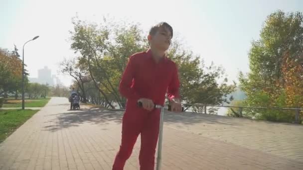 Lindo chico con camisa roja y pantalones montando una scooter en el parque. El niño pasa activamente tiempo al aire libre. Movimiento lento . — Vídeos de Stock