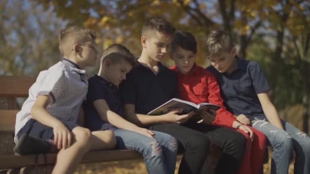 Cinco chicos sentados en el banco y montando una revista. Amigos pasan tiempo en gran compañía en un día soleado en el parque de otoño . — Vídeo de stock