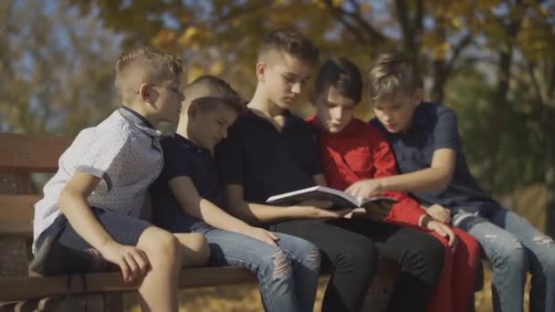 Compañía de chicos sentados en el banco y montando una revista. Amigos pasan tiempo montando revista en un día soleado en el parque de otoño . — Vídeo de stock