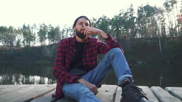 Бородатый юнг сидит возле удивительного озера и курит сигары — стоковое видео